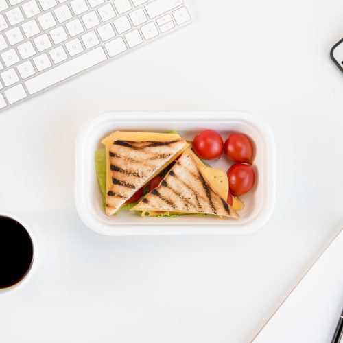 Top view of sandwiches with tomatoes in lunch box, notebook with pen, cup of coffee, smartphone and keyboard at workplace